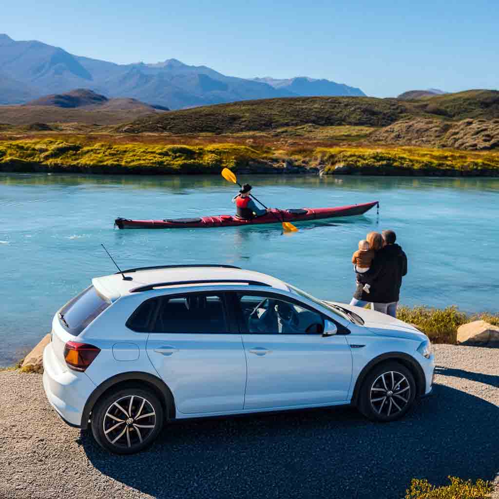Volkswagen Polo Track MSI MT de alquiler en San Carlos de Bariloche, estacionado a orillas del río Limay mientras una pareja observa un kayak de travesía. Servicio Rent a Car de Diversidad. Turismo Activo & de Naturaleza.